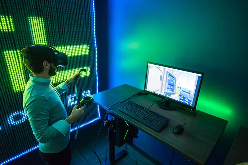 A person wearing a virtual reality headset holds two joysticks in hand as he stands in front of a monitor displaying his view in the virtual reality headset.