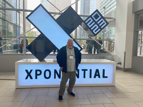 Bill Adams stands in front of a sculpture of a big X with a small cube that reads 2023, that is sitting on top of a box that reads XPONENTIAL at the AUVSI XPONENTIAL Conference and trade show in Denver Colorado in May 2023.