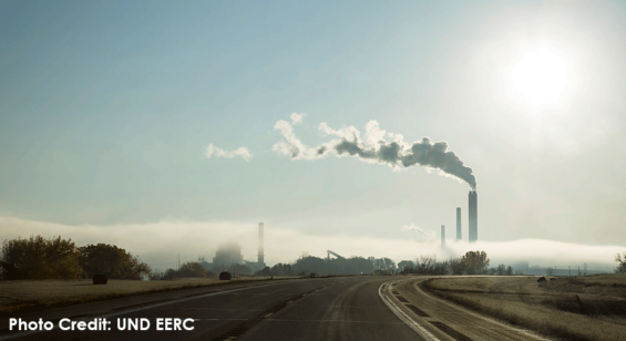 Behind the bend in the road lays Coal Creek Electricity Plant with 4 smoke stacks in the skyline. The tallest stack billows out smoke as the sun shines brightly behind it. A layer of fog is sitting just over the plant.