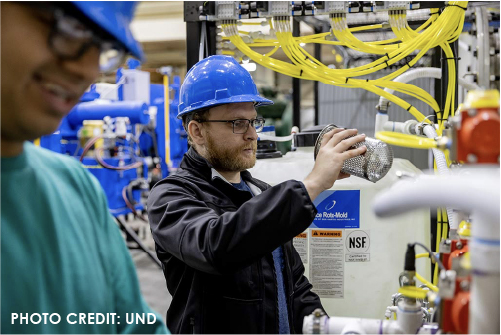 In a large warehouse, a man wearing a hard hat and glasses holding a metal cylinder one foot away from his face looking into it. The man is framed by another worker and chemical/liquid tubes and storage containers.