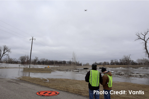 Two individuals stand in bright safety vests in front of the flooded Red River in Grand Forks, North Dakota. A drone hovers above them and the river is in the background.