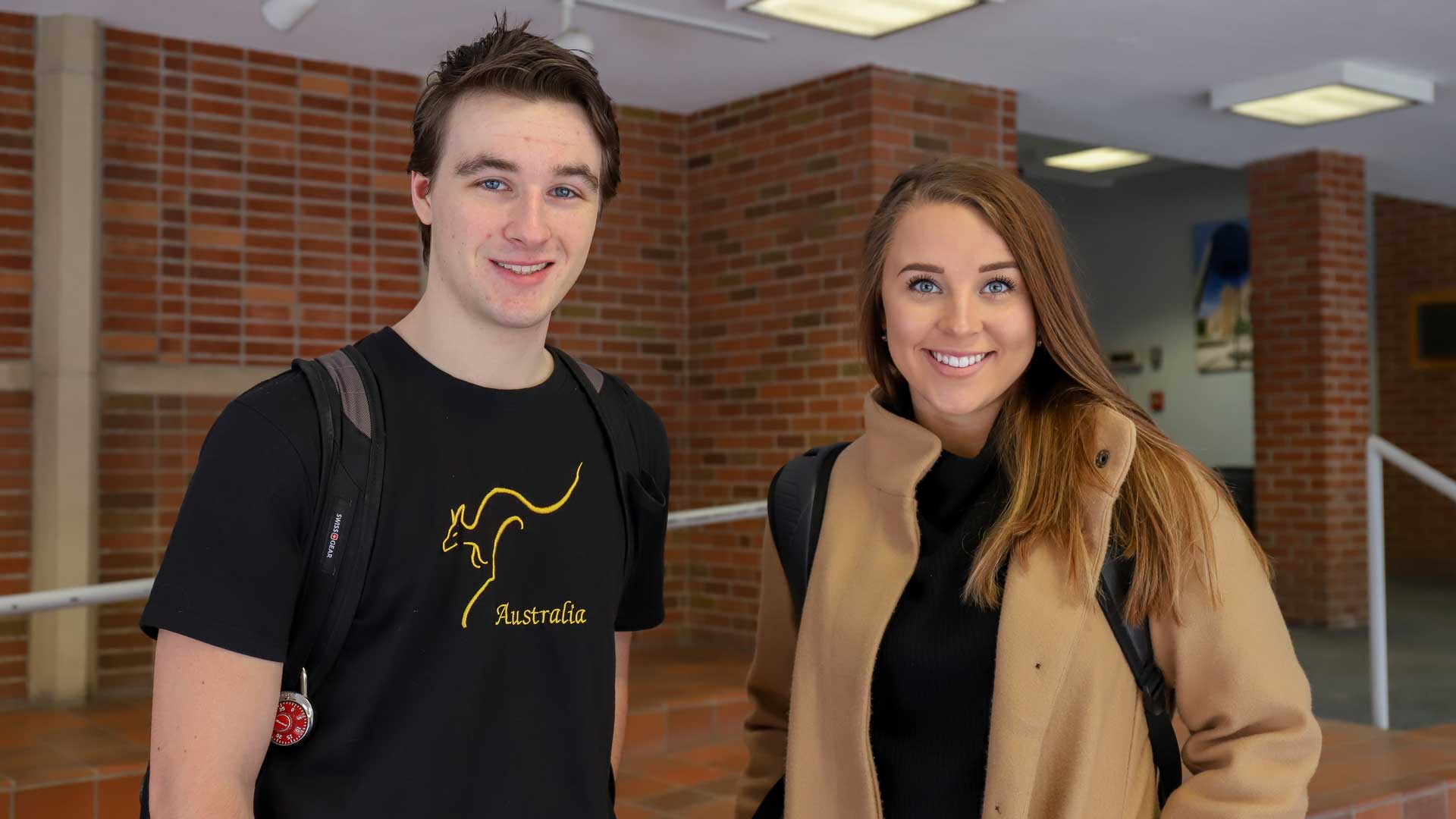 Two students stand together smiling in a college university building with a brick wall behind them.