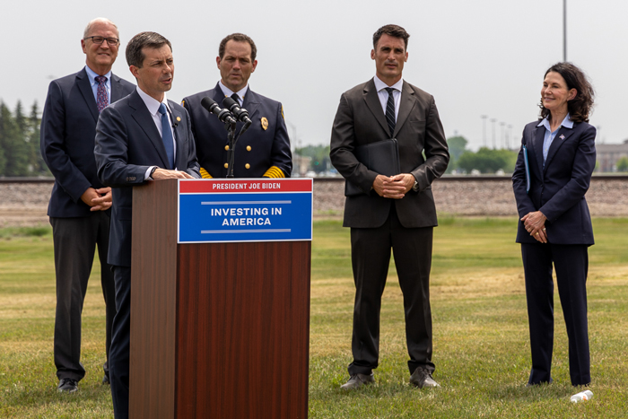 US Secretary of Transportation Pete Buttigieg stands at a podium that has a sign stating President Joe Biden Investing in America as he announces a $30 million investment for an underpass for 42nd St, Demers Ave, and the railroad tracks at that intersection. Behind Buttigieg is US Senator Kevin Cramer, Grand Forks Fire Chief Gary Lorenz, Mayor Brandon Bochenski, and Lieutenant Governor Tammy Miller.