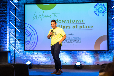 Brandon Baumbach stands on a stage presenting at the Pillars of Place conference in Downtown Grand Forks, North Dakota.