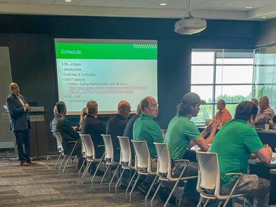 A group of people sit at tables that are positioned in a u facing a presenter who stands by a screen that reads SCHEDULE, 3-4 PM, INTRODUCTIONS, OVERVIEW OF CURRICULUM" at the UAS curriculum meeting at the University of North Dakota.