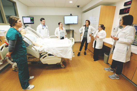 A group of professionals in the medical industry stand around a medical bed talking.
