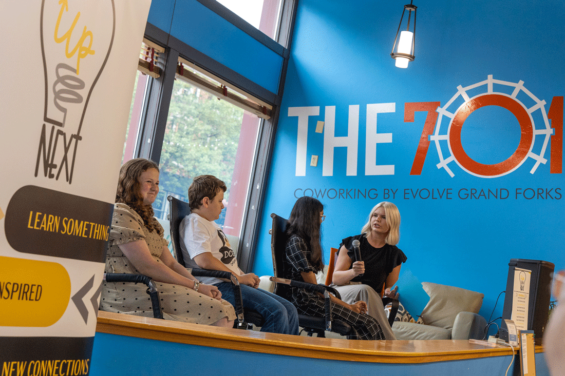 Four people sit on a stage presenting with The 701 coworking logo behind them on the wall and the UpNext sign in the front of them.
