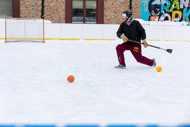 A man playing broom ball in the Grand Forks Town Square ice rink in the winter.