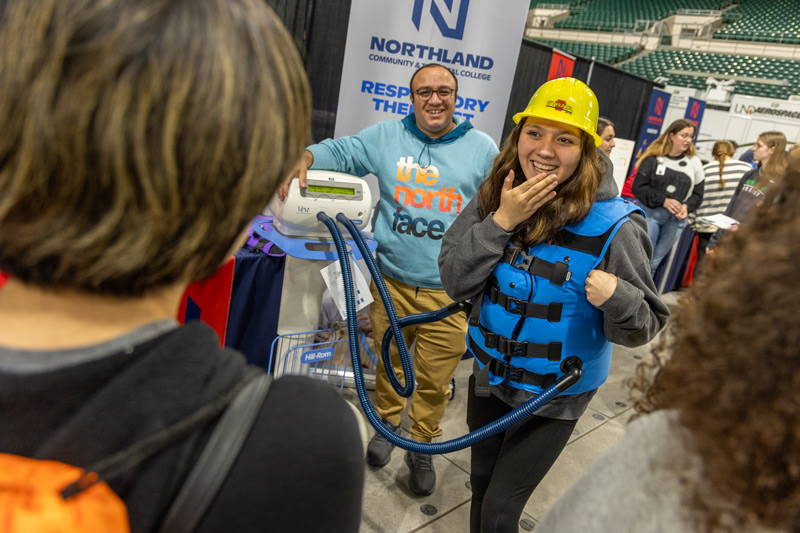 A student at the Northern Valley Career Expo smiles while wearing a medical vest that is hooked up to a machine while she looks at her friends standing by. 