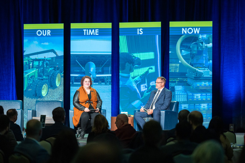 EDC Director of Workforce Development Becca Cruger and President/CEO Keith Lund sit on stage in front of electronic panels reading, "Our Time Is Now" at the EDC Annual Meeting on March 26th, in front of a crowd of people.