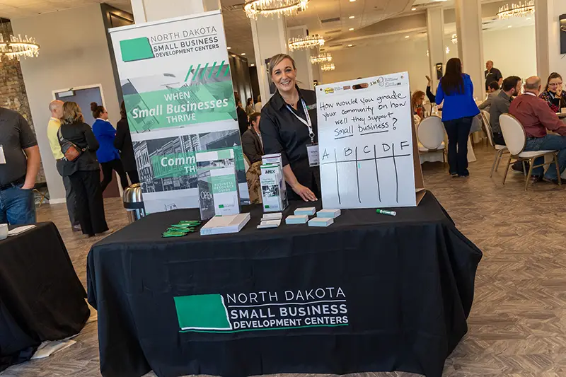 A woman staffs a North Dakota Small Business Development Centers table with informational posters and a community grading chart at a conference.