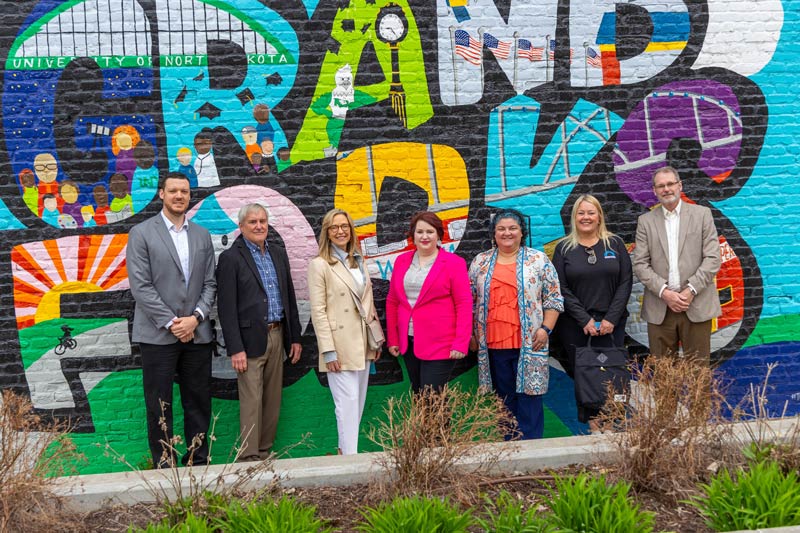 A group of individuals stand together in front of the Grand Forks mural in town square of downtown Grand Forks.