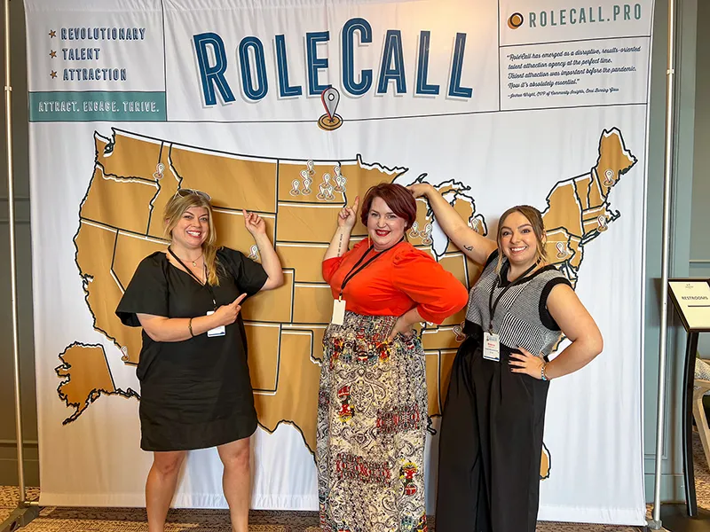 Becca Cruger and Brenna Mathiason stand in front of a banner that reads RoleCall with a map of the USA alongside a RoleCall employee.