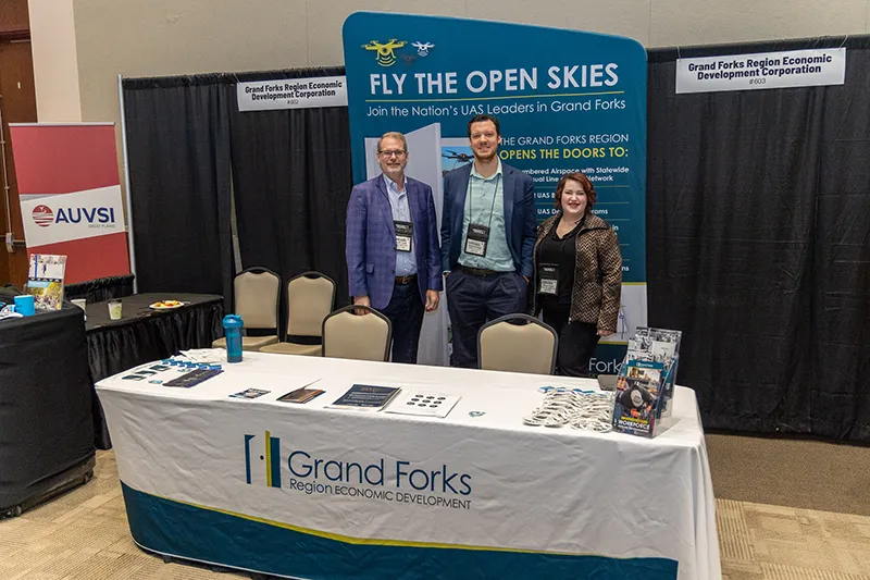 EDC Staff stand in front of an expo booth banner that reads Fly the Open Skies and a table with the Grand Forks Region EDC logo on it.