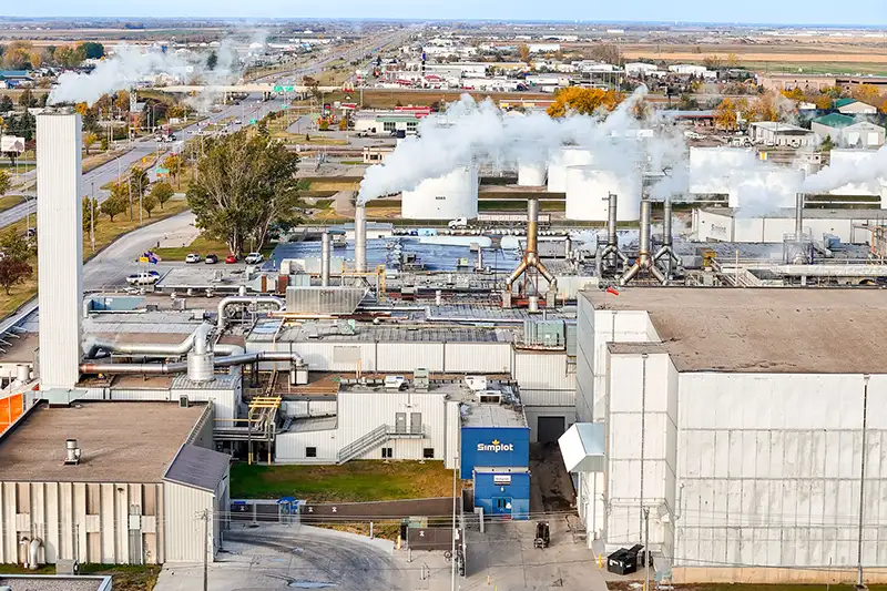 An aerial drone view of the J.R. Simplot food processing plant in Grand Forks, showing the large white high-bay factory buildings, a tall smokestack on the left, and multiple steam plumes rising from rooftop vents and processing units, with a blue Simplot-branded loading dock visible at center, surrounded by parking areas, an adjacent highway, and the broader cityscape and autumn-colored countryside stretching to the horizon.