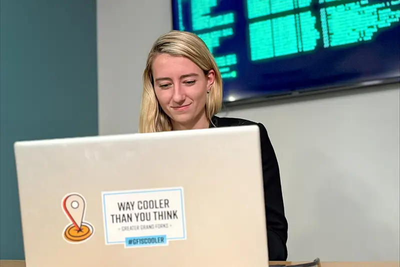 A young woman with shoulder-length blonde hair smiles softly as she works on a silver laptop covered with Greater Grand Forks "Way Cooler Than You Think" and "#GFisCooler" stickers, with a large wall-mounted monitor displaying lines of green and teal programming code in the background.