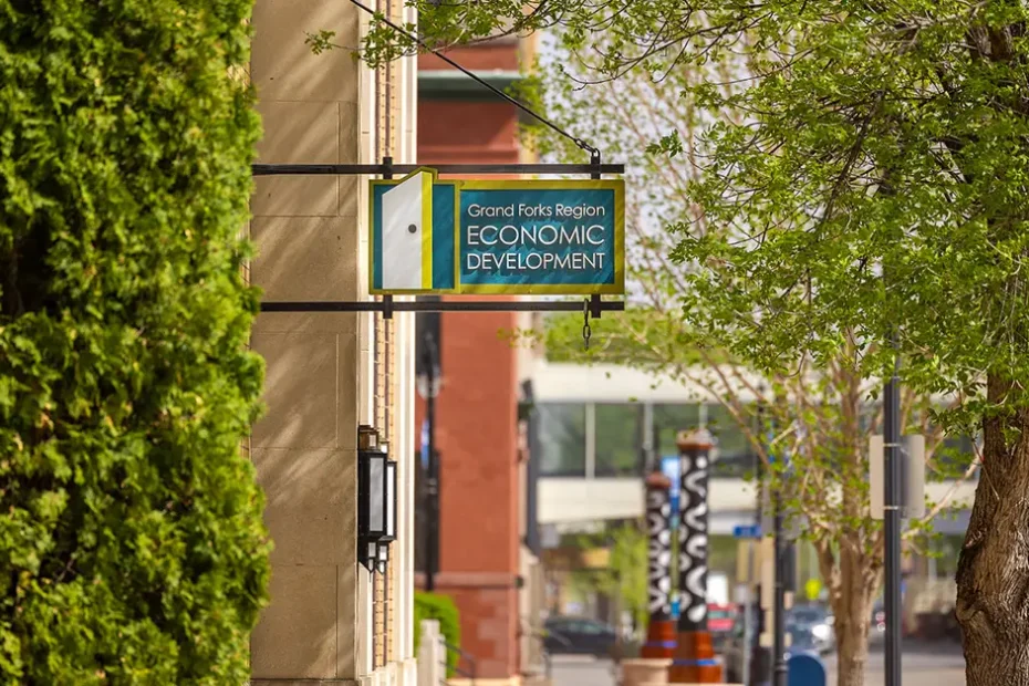 The Grand Forks Region Economic Development Corporation sign hanging from a building exterior on a tree-lined downtown street on a sunny spring day.