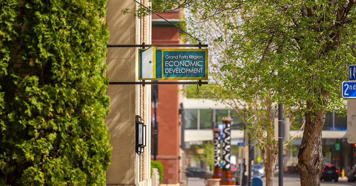 The Grand Forks Region Economic Development Corporation sign hanging from a building exterior on a tree-lined downtown street on a sunny spring day.
