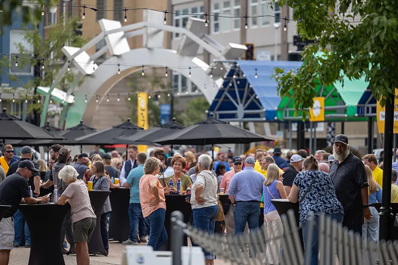 Community members gather at cocktail tables socializing in town square, with the paddle wheel prominently displayed behind them.