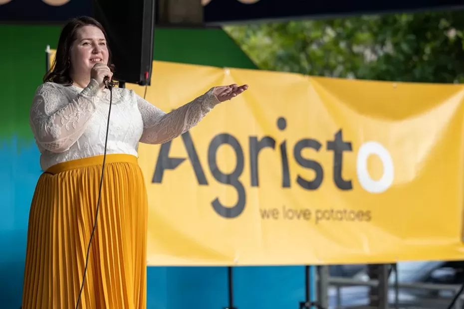 Becca Cruger stands on a stage holding a microphone in front of a banner of the Agristo logo at the community welcome celebration.