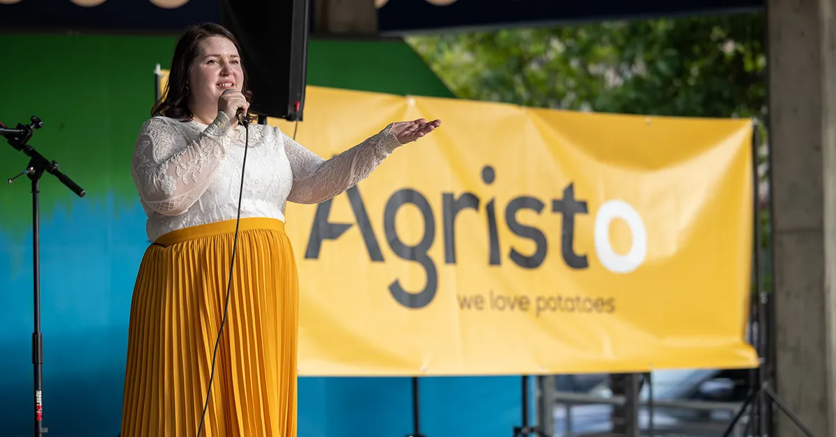 Becca Cruger stands on a stage holding a microphone in front of a banner of the Agristo logo at the community welcome celebration.