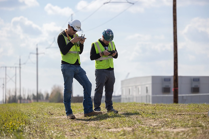 Two men wearing reflective vests, they are flying drones.