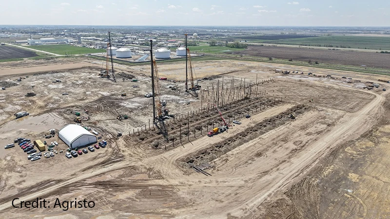 Aerial view of the Agristo construction site showing rows of steel foundation columns, cranes, and heavy equipment on a large cleared plot, with the Grand Forks skyline visible in the distance.