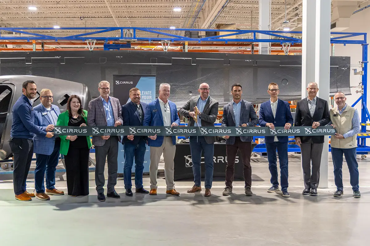 A line of business and community leaders cutting a ribbon at a Cirrus Aircraft facility, with an aircraft fuselage in the background.