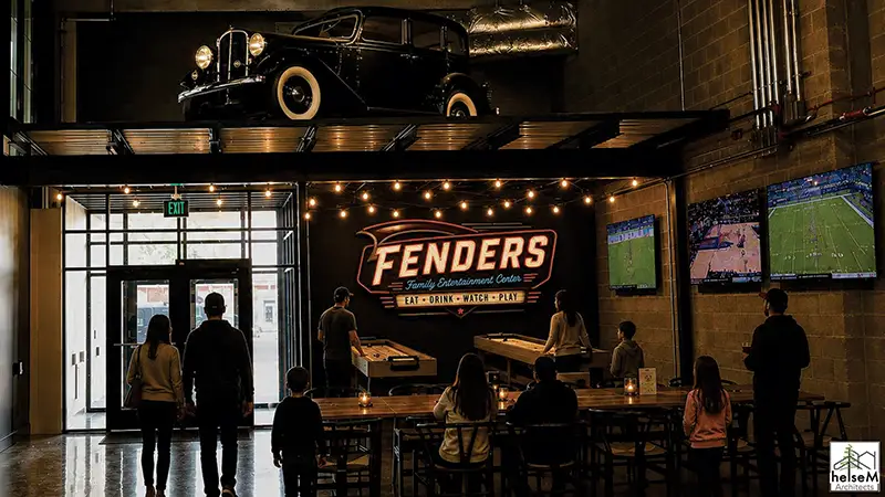 Interior of Fenders Family Entertainment Center, with a lit Fenders sign, shuffleboard tables, wall-mounted TVs showing sports, and patrons gathered at tables beneath a vintage black car displayed on a mezzanine.