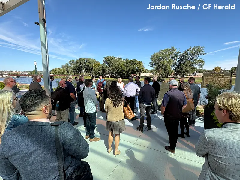 Grand Forks leaders standing on a riverfront plaza in Sioux Falls, South Dakota, listening to a presentation, with the water, trees, and a Seney Island historical marker in the background. Photo credit: Jordan Rusche / GF Herald.