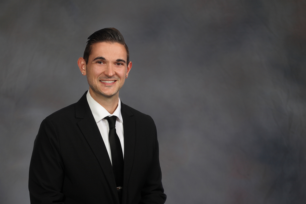 mark maliskey is smiling in front of a photography backdrop wearing a dark suit and tie