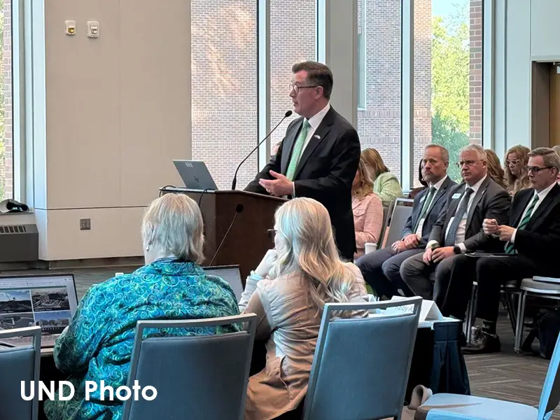 A man in a dark suit and green tie speaks at a podium during a legislative committee meeting at UND, while attendees sit in the foreground and lawmakers sit at right. Photo credit: UND Photo.