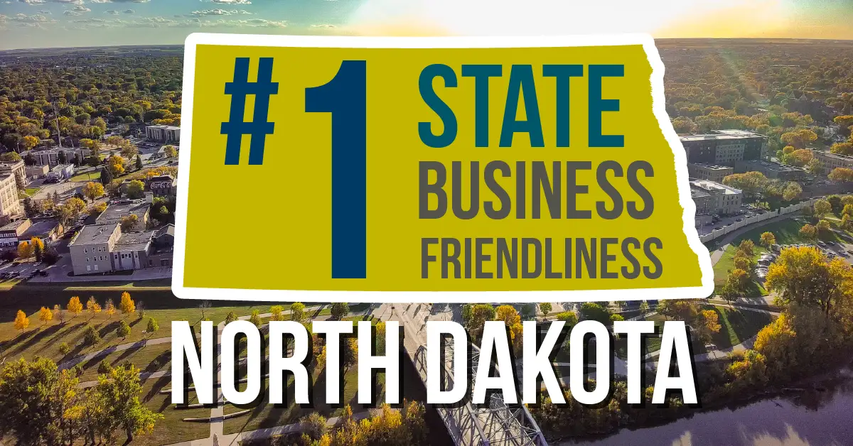 Graphic reading '#1 State Business Friendliness, North Dakota' over an aerial photo of downtown Grand Forks along the Red River in fall, with a pedestrian bridge crossing the water in the foreground.
