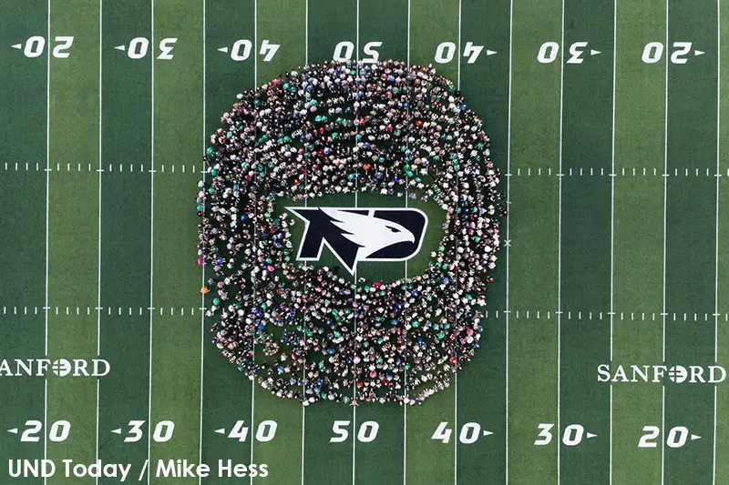 Overhead view of a large crowd of UND students and staff arranged in a ring around the Fighting Hawks logo at midfield of a football field. Photo credit: UND Today / Mike Hess.