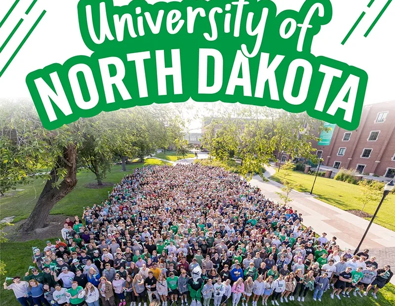 A massive group of University of North Dakota students, the Class of 2029, is gathered on a grassy quad for a class photo. The students are densely packed together, filling the center of the frame and forming a large block. Many are wearing green or white UND apparel. The photo is taken from a slightly elevated angle, showing a wide view of the group with campus buildings and trees visible on either side. The top of the image features the text "University of North Dakota" in a stylized, bold green font with a white outline.