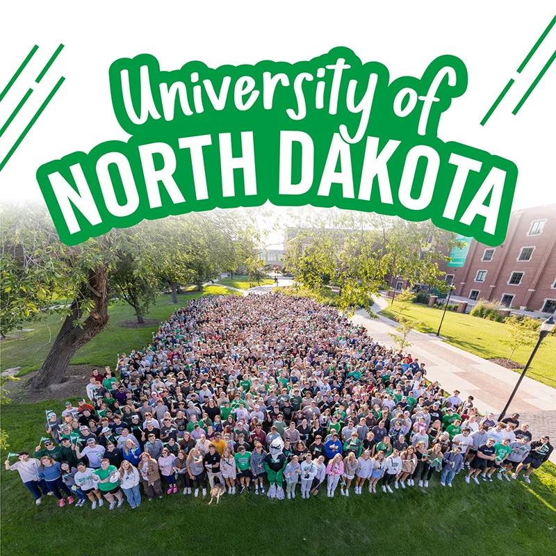 A massive group of University of North Dakota students, the Class of 2029, is gathered on a grassy quad for a class photo. The students are densely packed together, filling the center of the frame and forming a large block. Many are wearing green or white UND apparel. The photo is taken from a slightly elevated angle, showing a wide view of the group with campus buildings and trees visible on either side. The top of the image features the text "University of North Dakota" in a stylized, bold green font with a white outline.